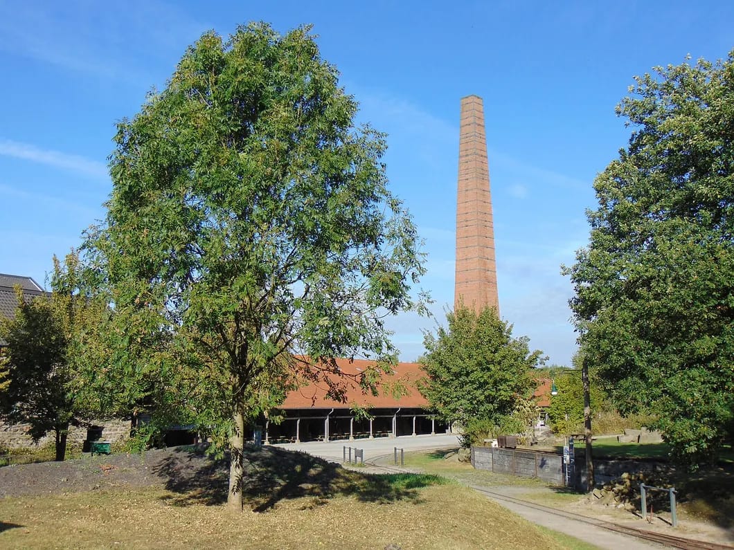 Hoher Ziegelschornstein hinter einer langen Halle mit rotem Ziegeldach, davor eine Wiese mit großen Laubbäumen.