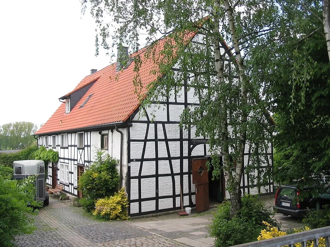 Fachwerkhaus mit rotem Ziegeldach und Schleppgaube, seitlich von Birken und Sträuchern gerahmt, davor eine gepflasterte Hofzufahrt.