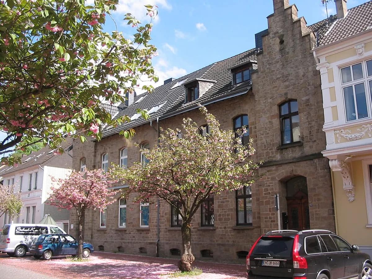 Zeile historischer Bruchsteinhäuser mit Staffelgiebel an einer Straße, davor zwei blühende Zierkirschen an einem sonnigen Frühlingstag.