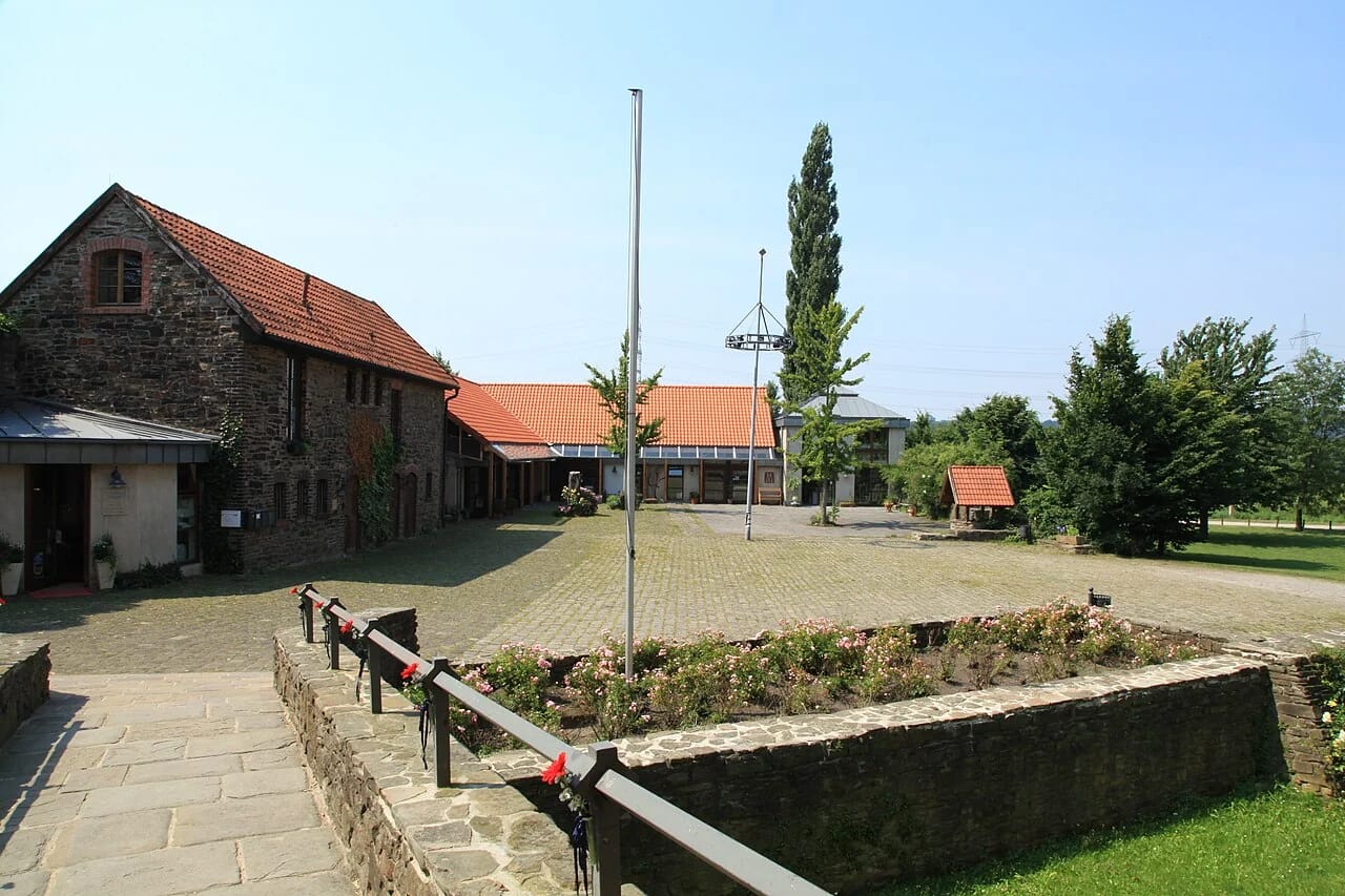 Gepflasterter Innenhof von Haus Herbede mit Bruchsteingebäuden unter roten Ziegeldächern und einem von einer Mauer gefassten Rosenbeet in der Mitte.