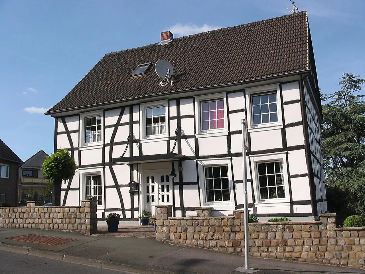 Zweigeschossiges Fachwerkhaus mit weiß verputzten Gefachen und dunklem Ziegeldach, davor eine Bruchsteinmauer und ein schmaler Vorgarten.