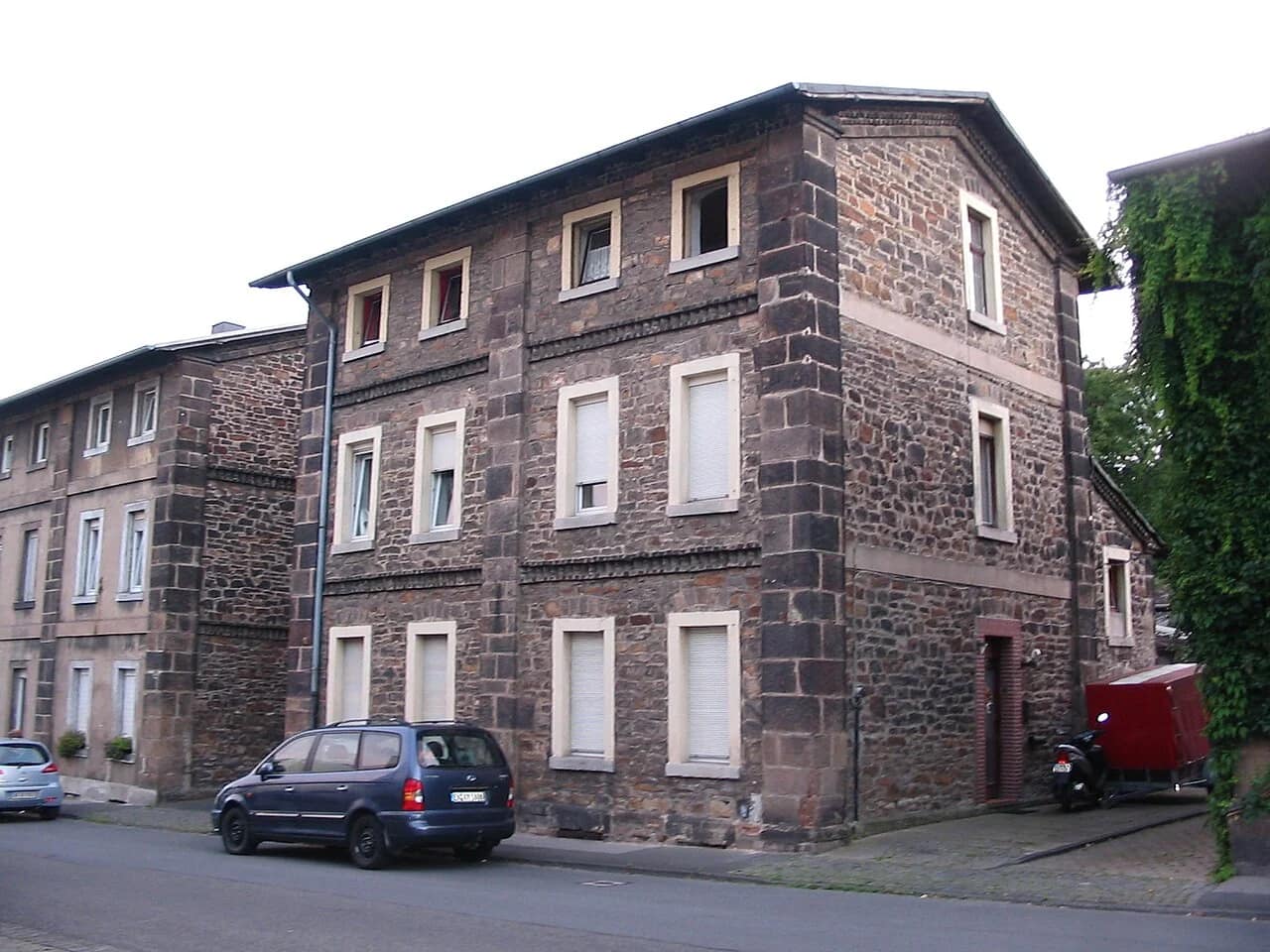 Dreigeschossiges Bruchsteinhaus mit hellen Fensterfaschen an einer abschüssigen Straße, links schließt eine gleichartige Häuserzeile an.
