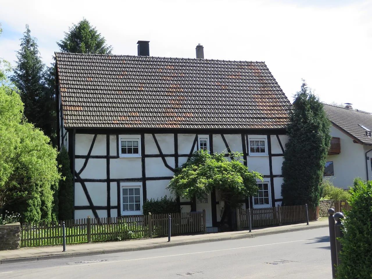 Langgestrecktes Fachwerkhaus mit steilem Schieferdach an einer Straße, die Fassade teilweise von einem blühenden Kletterstrauch überwachsen.
