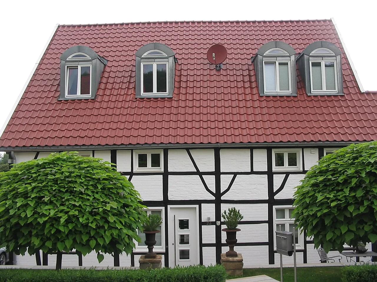 Weiß gestrichenes Fachwerkhaus mit rotem Ziegeldach und vier Dachgauben, davor zwei kugelförmig geschnittene Bäume und Kübelpflanzen am Eingang.