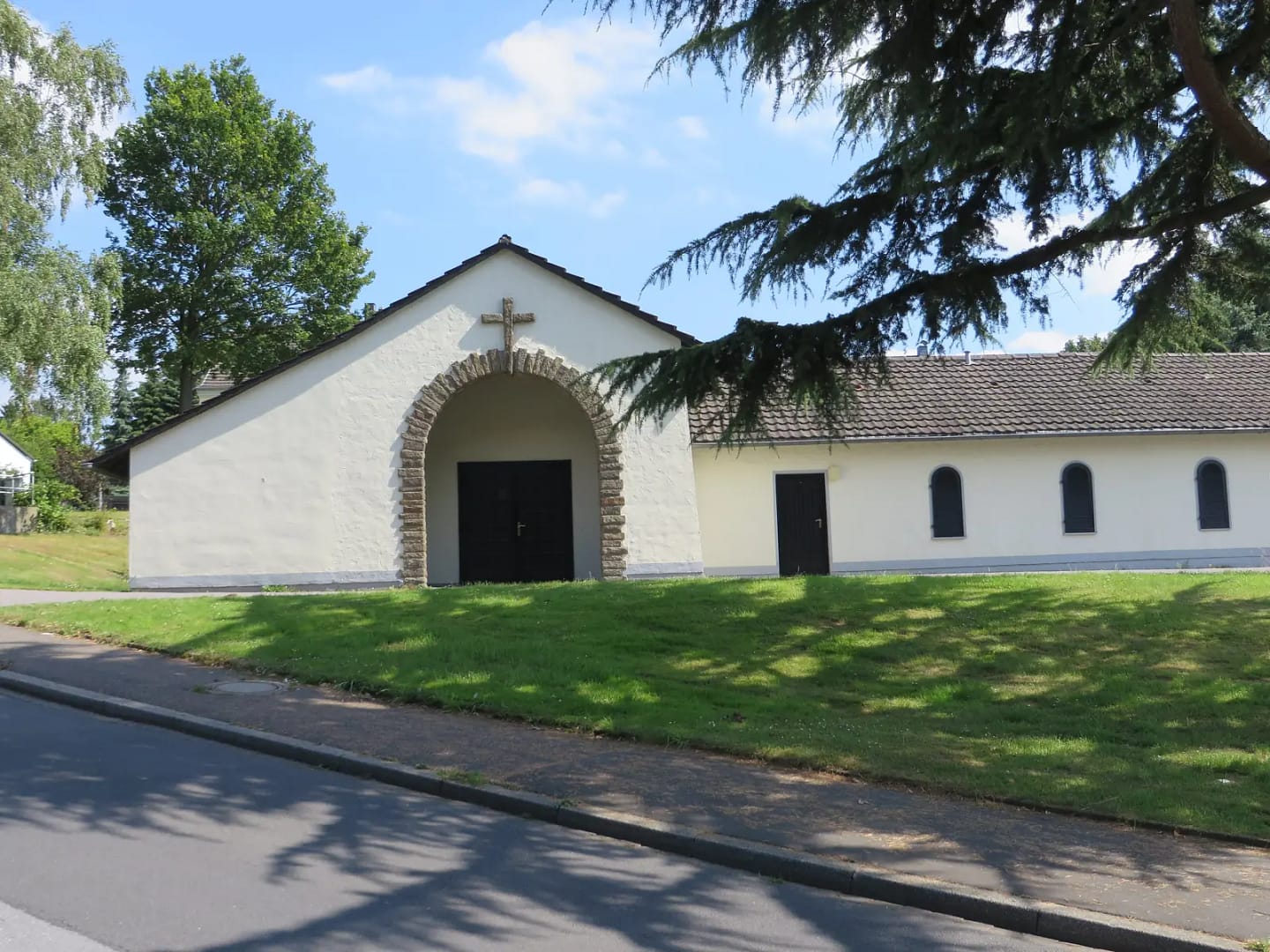 Schlichte weiß verputzte Kapelle mit Rundbogenportal und Steinkreuz über dem Giebel, davor eine Rasenböschung im Schatten einer Zeder.