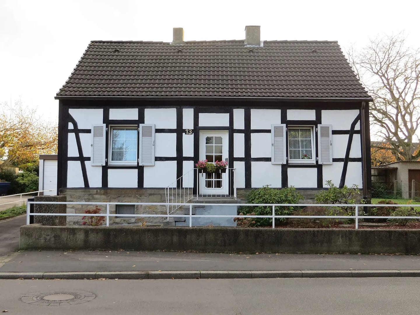 Breites Fachwerkhaus mit hellen Klappläden und Blumenkasten über der Haustür, davor eine Stützmauer aus Bruchstein mit weißem Geländer.