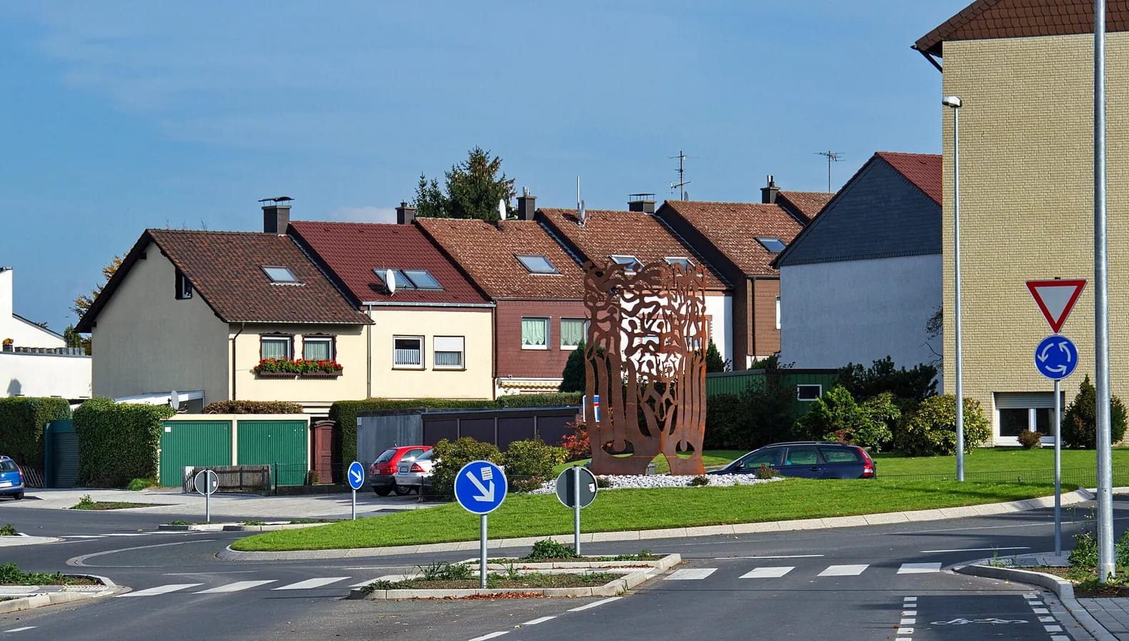 Reihe von Ein- und Zweifamilienhäusern mit Sattel- und Krüppelwalmdächern hinter Hecken und Garagen, davor ein Kreisverkehr mit einer rostroten Stahlskulptur.