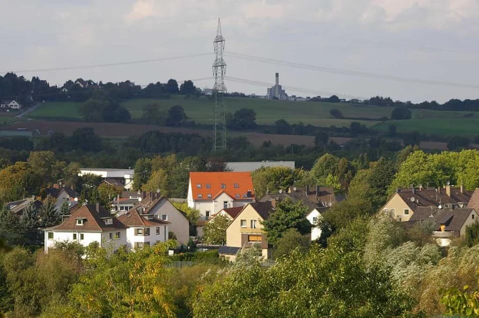 Blick über die Dächer von Ein- und Mehrfamilienhäusern zwischen hohen Bäumen, dahinter Felder, ein Strommast und ein bewaldeter Höhenzug.